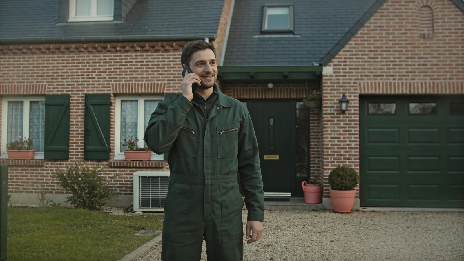 Un chauffagiste en combinaison verte téléphone devant une maison en brique avec garage, sur une allée de gravier