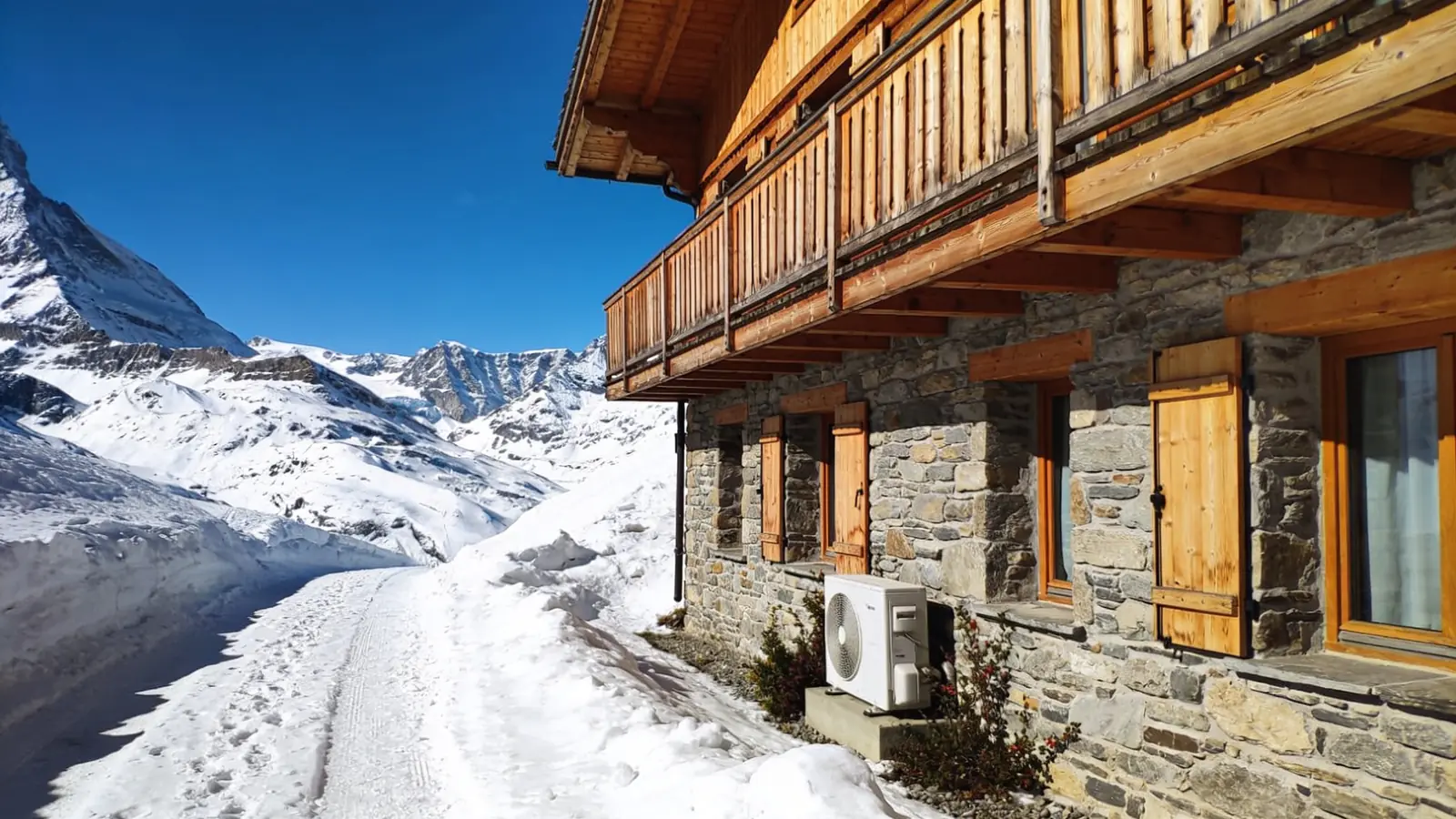 Maison de montagne en pierre avec étage et balcon en mélèze, sentier enneigé et sommets blancs sous un ciel bleu, une pompe à chaleur posée contre le soubassement