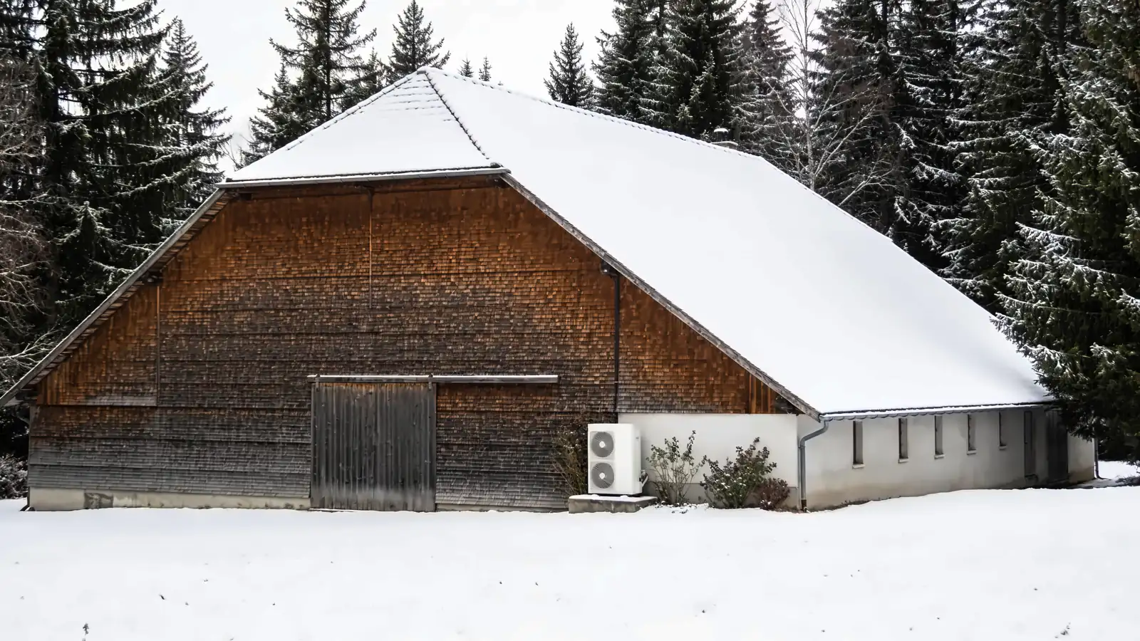 Grande ferme comtoise au pignon couvert de tavaillons et toit enneigé, sapins et champ sous la neige en hiver, une pompe à chaleur posée contre la partie maçonnée