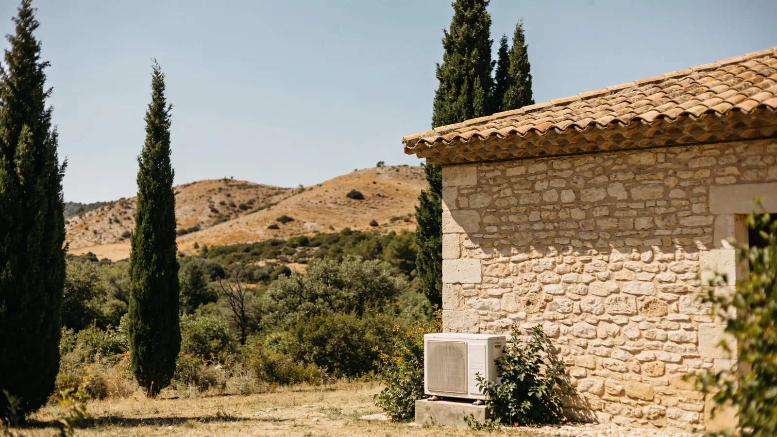 Mas gardois en pierre claire au toit de tuiles canal, cyprès, garrigue et collines sèches en plein été, une pompe à chaleur posée sur un socle contre le mur