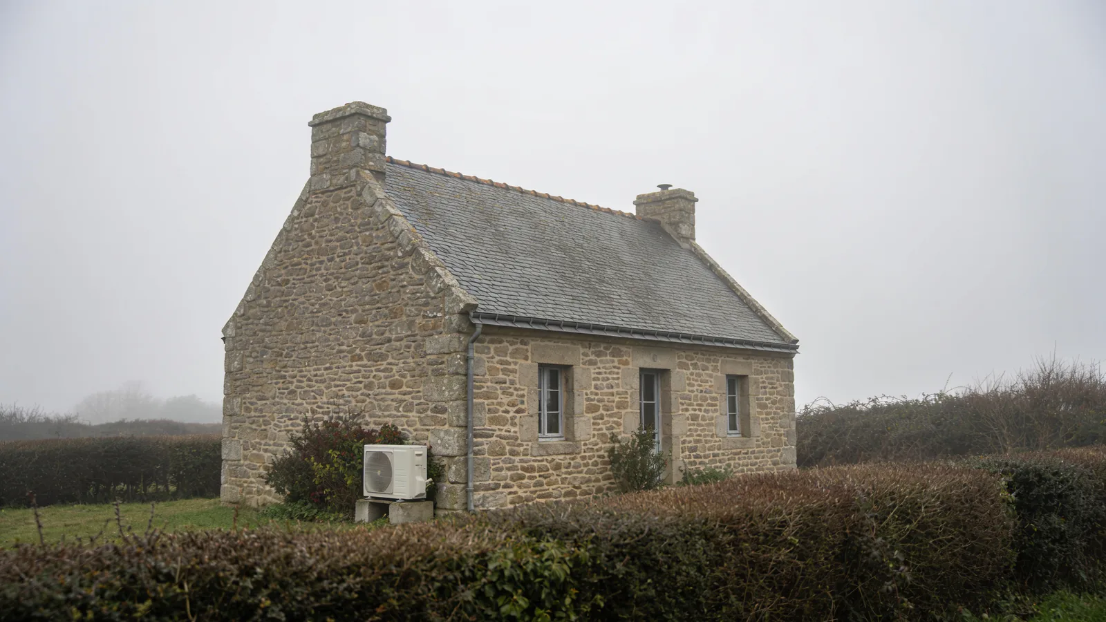 Maison du Cotentin en granit au toit d'ardoise derrière les haies du bocage, par une journée d'hiver grise et brumeuse, une pompe à chaleur posée contre le mur