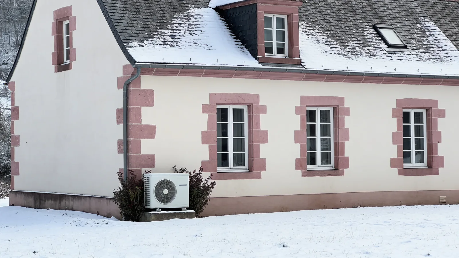 Maison mosellane enduite de blanc aux encadrements de grès rose et toit d'ardoise enneigé, neige au sol en hiver, une pompe à chaleur posée contre la façade