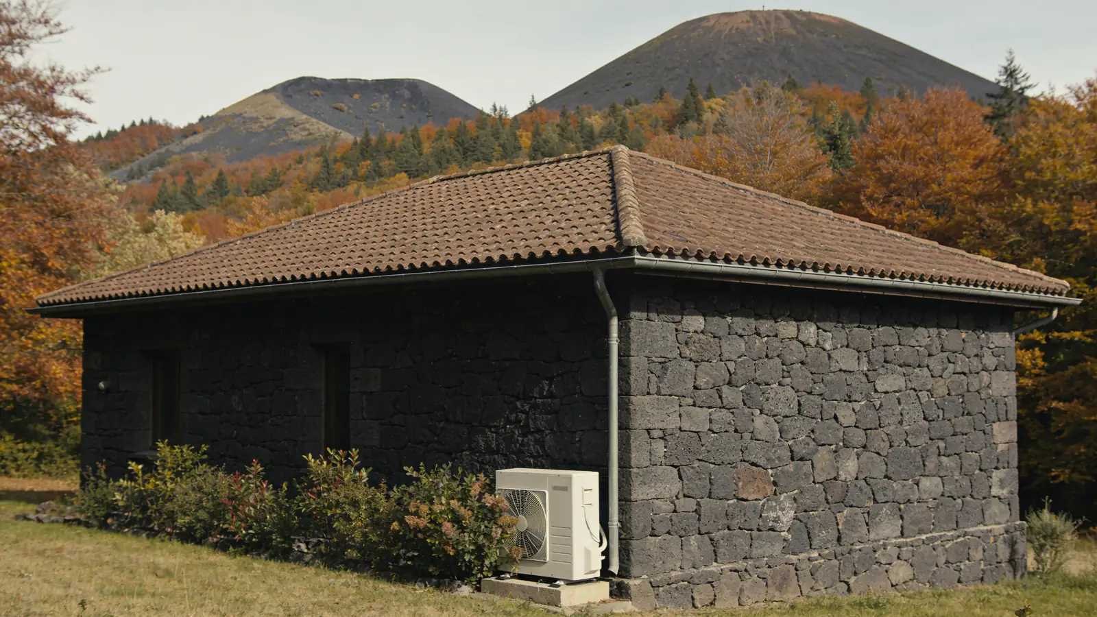 Maison auvergnate en pierre volcanique gris sombre au toit de tuiles brunes, puys boisés en arrière-plan en automne, une pompe à chaleur posée contre le mur