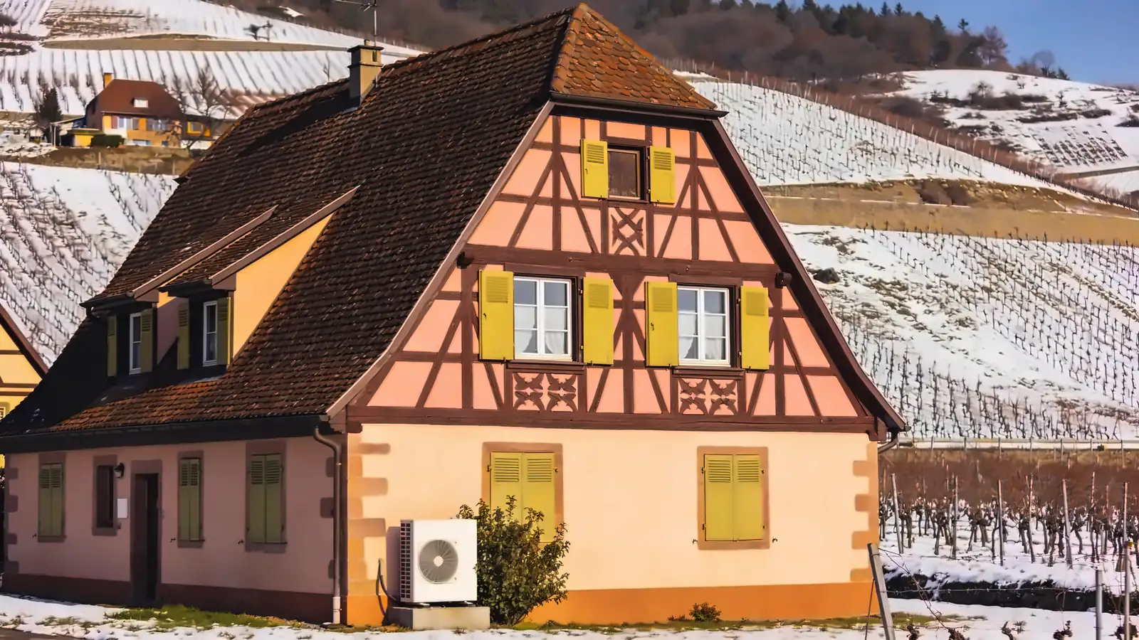 Maison alsacienne colorée à colombages et volets verts dans un village viticole, vignes enneigées sur les coteaux en hiver, une pompe à chaleur posée contre la façade