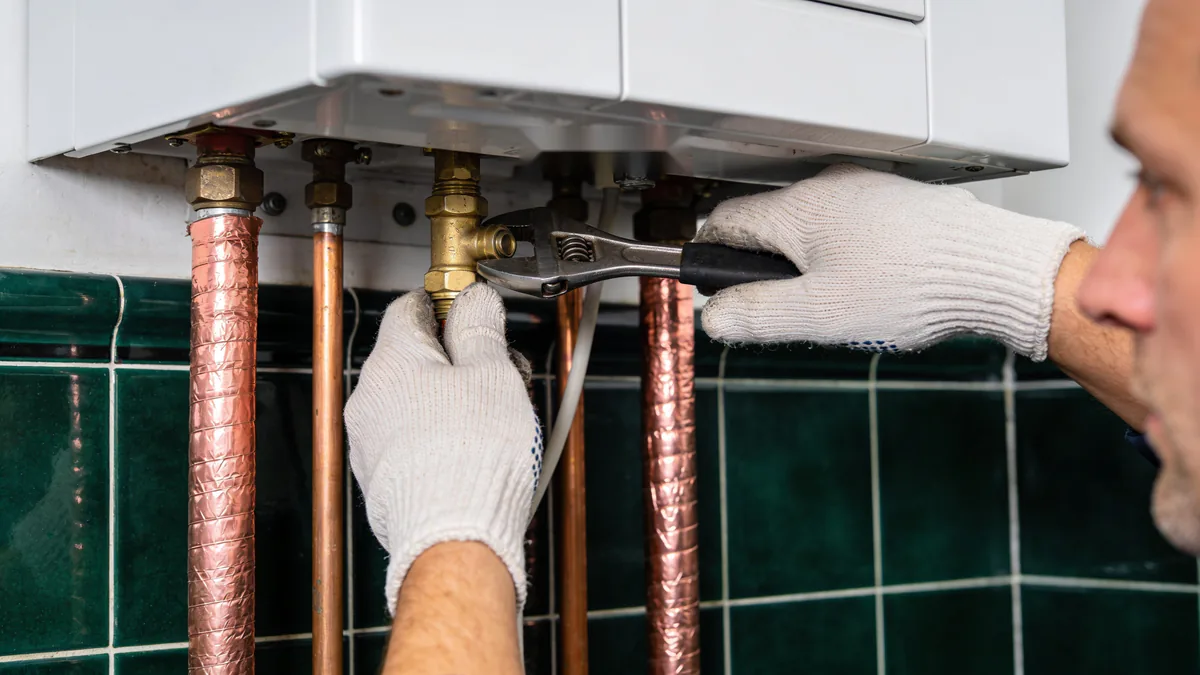 Des mains gantées resserrent à la clé un raccord en laiton sous le module intérieur d'une pompe à chaleur, entre des tuyaux de cuivre isolés