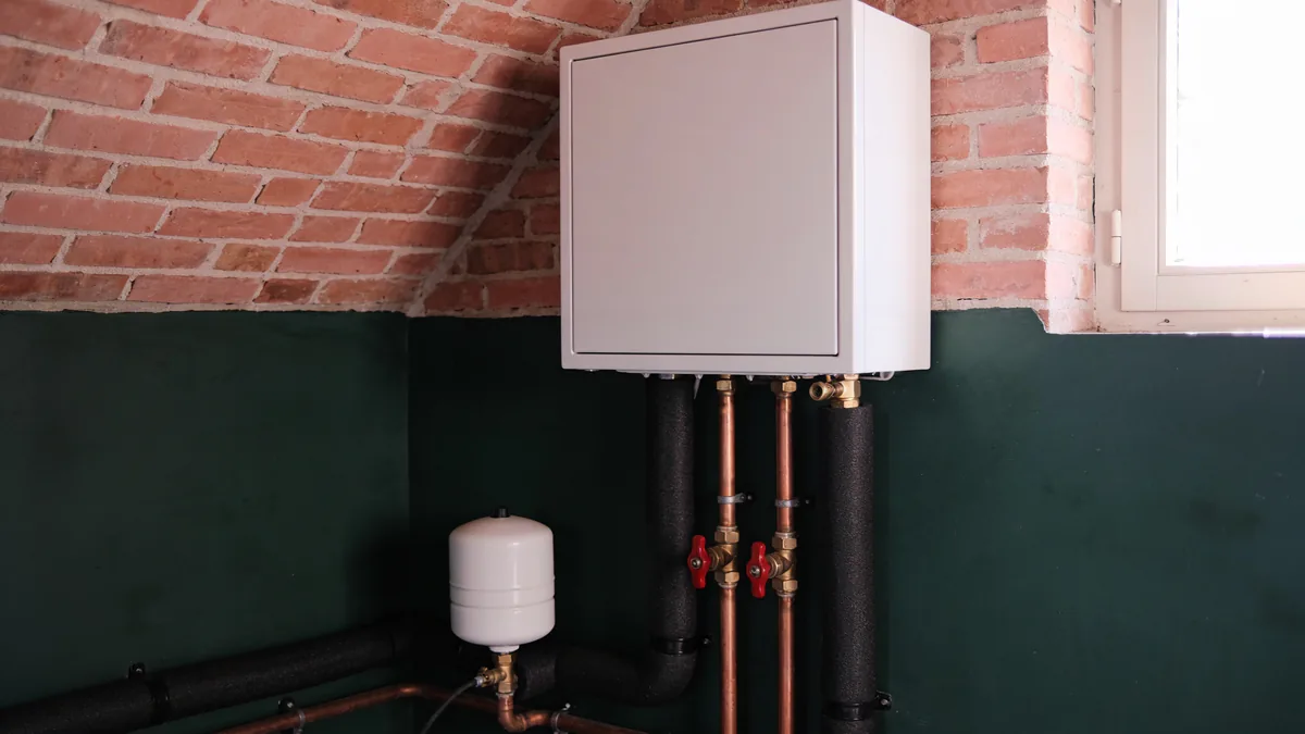Module hydraulique blanc fixé au mur d'une chaufferie aux murs de brique et vert foncé, avec ses tuyaux de cuivre isolés de mousse noire, des vannes à poignée rouge et un vase d'expansion blanc