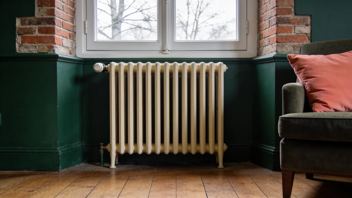 Radiateur en fonte peint couleur crème avec robinet thermostatique, sous une fenêtre à petits carreaux, dans un salon aux murs vert foncé et au parquet ancien