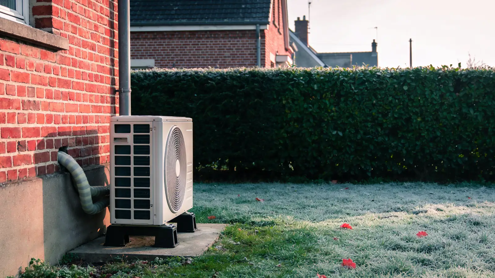 Unité extérieure blanche d'une pompe à chaleur air/eau posée sur des supports noirs et une petite dalle, contre le mur en brique rouge d'une maison du Nord, pelouse givrée et haie verte un matin d'hiver