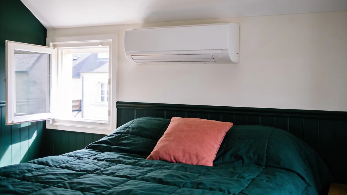 Unité murale blanche d'une pompe à chaleur air/air fixée au-dessus d'un lit au couvre-lit vert, près d'une fenêtre ouverte